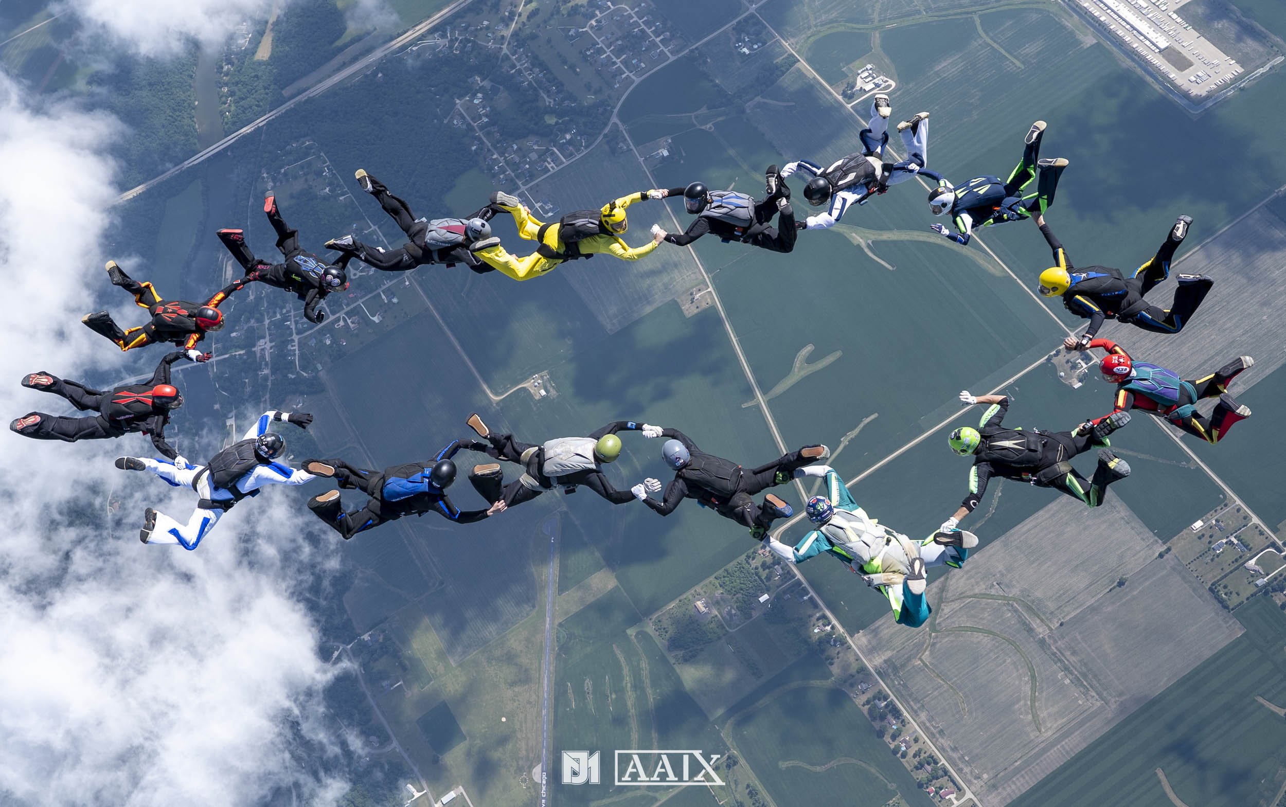 Group of skydivers forming a circle while free-falling above farmland and clouds.
