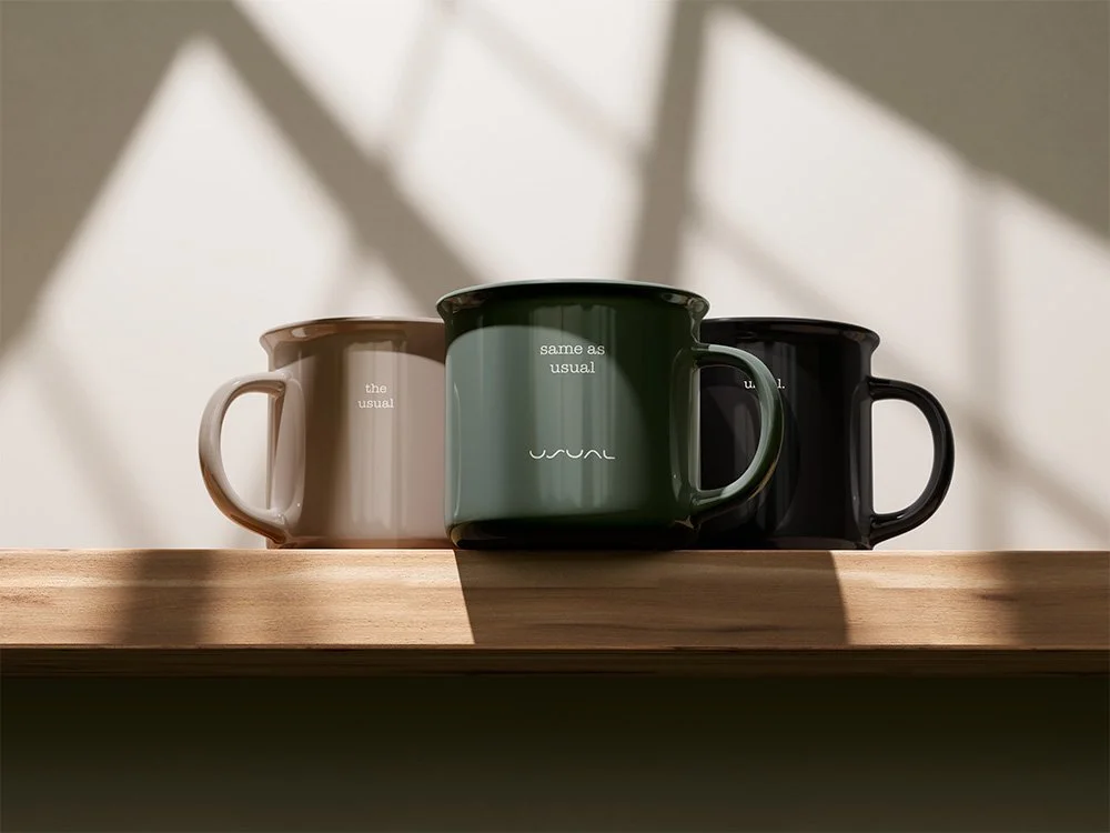 Three ceramic coffee mugs in neutral tones sit side by side on a wooden shelf in soft natural light. Each mug features minimal text and the understated “Usual” logo, emphasising familiarity, routine, and calm repetition.