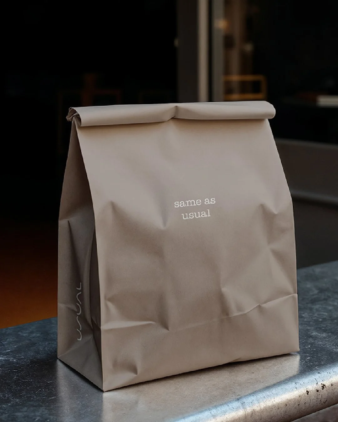 A neutral-toned paper takeaway bag placed on a café counter, folded at the top, with small white text reading “same as usual” on the front and a minimal logo printed on the side.