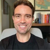 A smiling man with dark hair, wearing a black shirt, sitting in a room with bookshelves and a map in the background.