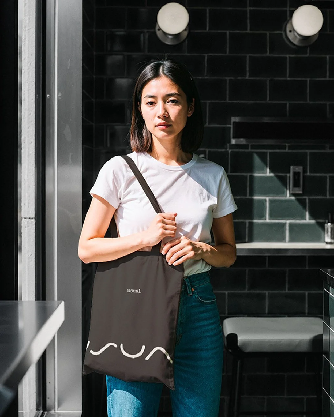 Person standing in a minimalist café interior holding a dark tote bag featuring The Usual logo, lit by natural window light.