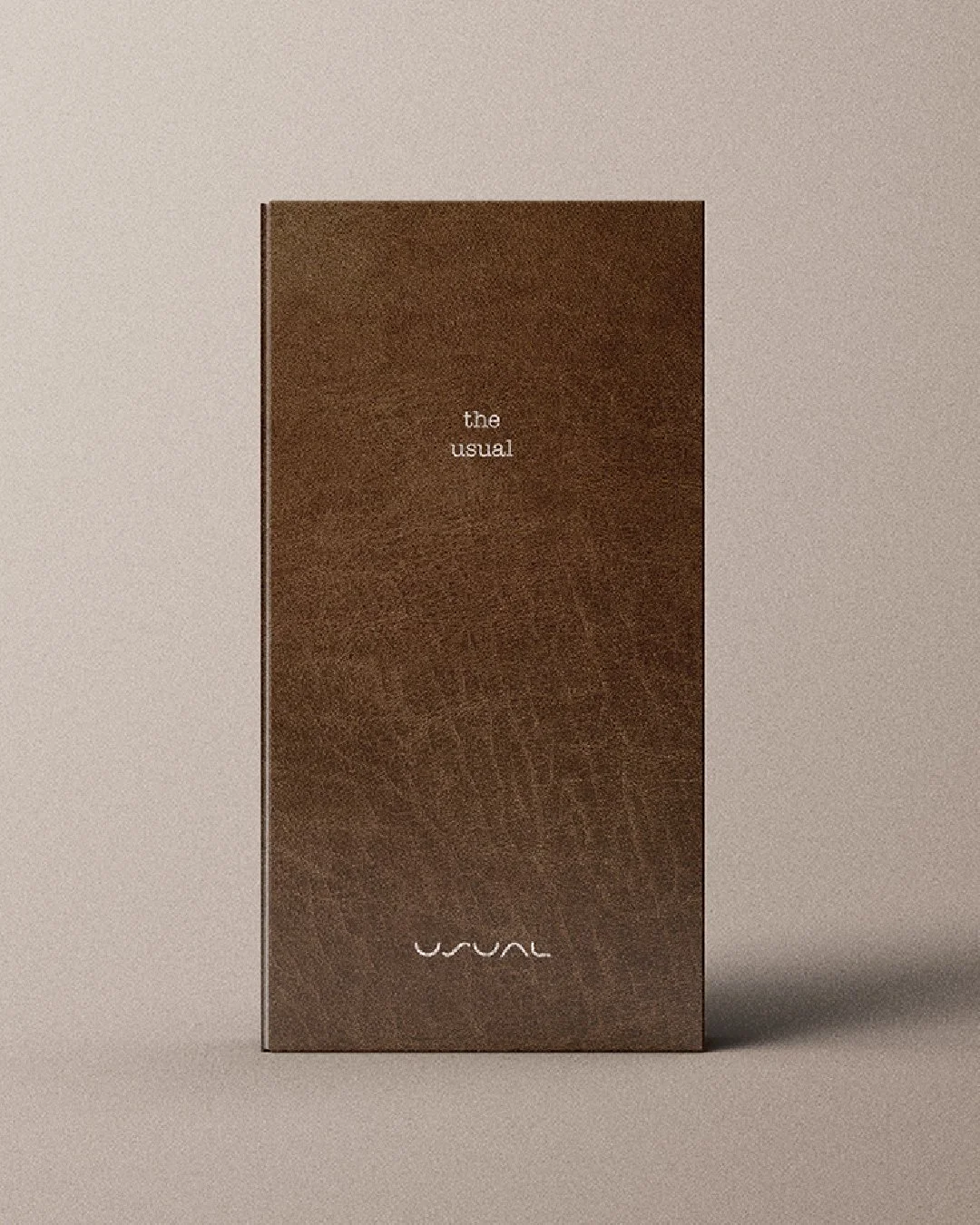 A tall, brown leather-textured café menu standing upright against a neutral background. The front cover features small white text reading “the usual” near the centre and a minimal logo at the bottom.