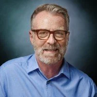 A middle-aged man with gray hair and a beard, wearing glasses and a blue shirt, smiling at the camera.