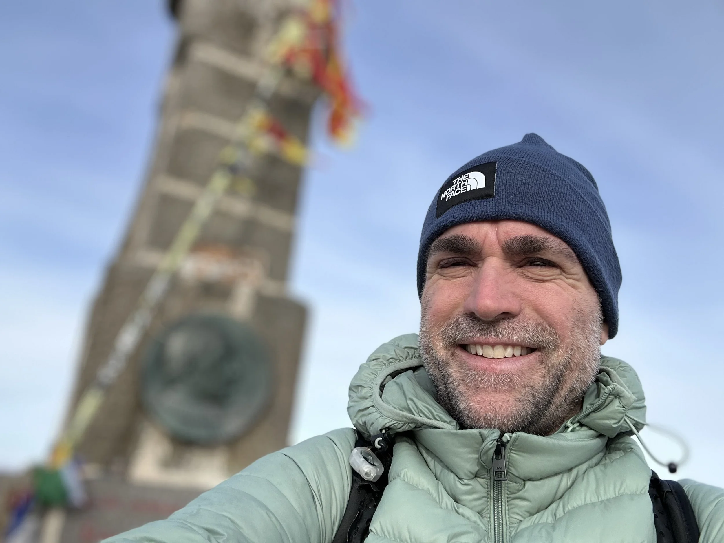 Close-up of a smiling man wearing a blue beanie and green jacket taking a selfie outdoors with a large stone cairn and a clear blue sky in the background.