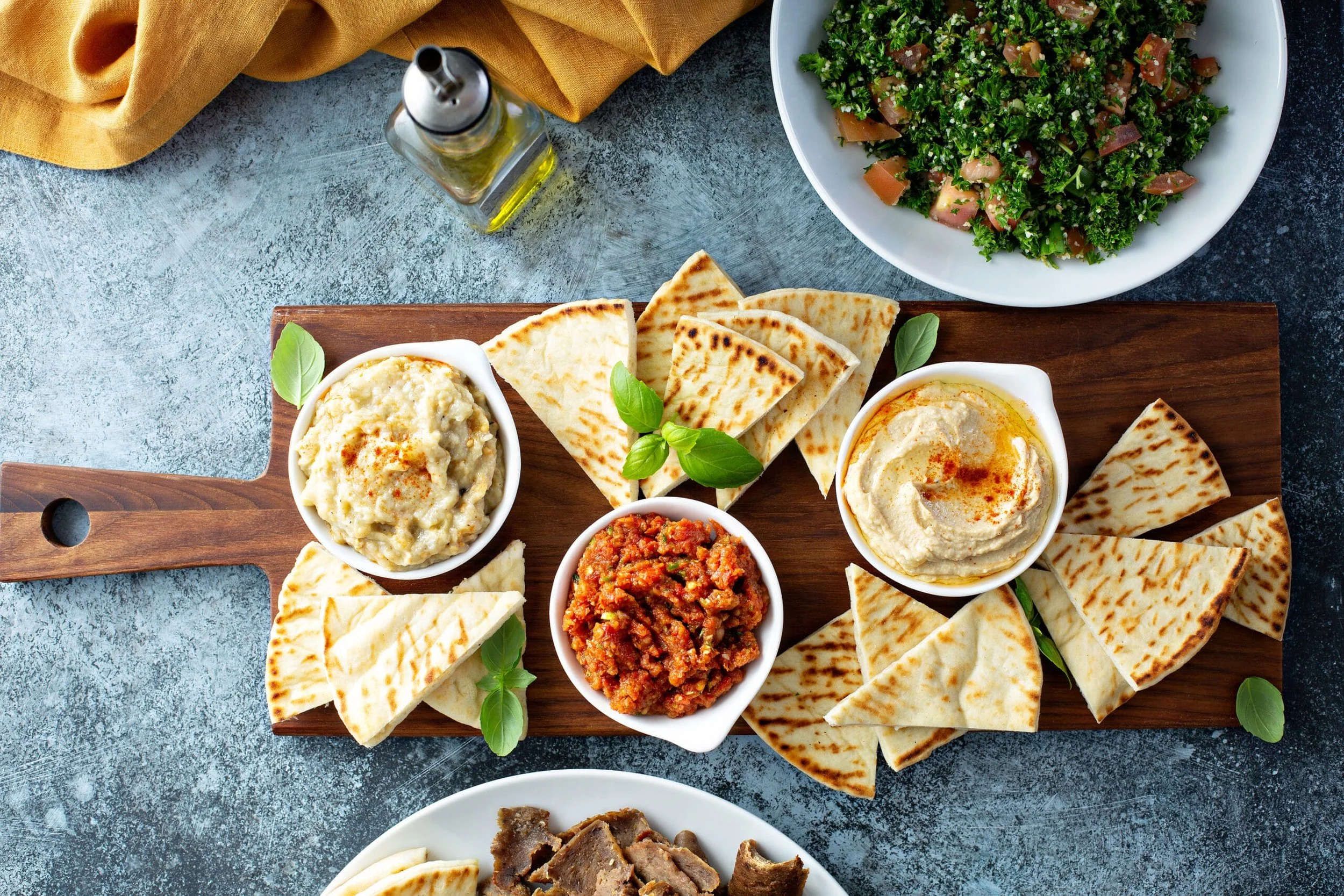 Assorted Middle Eastern dips and flatbreads on a wooden platter, with a bowl of tabbouleh salad and a small bottle of olive oil nearby.