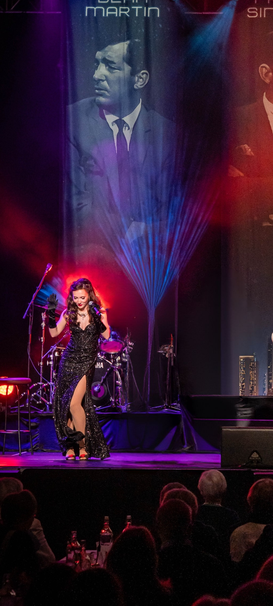 A woman in a black glittery dress with a high slit dancing on stage, with a large projected image of a man in a suit behind her, colorful stage lighting, and audience members in the foreground.