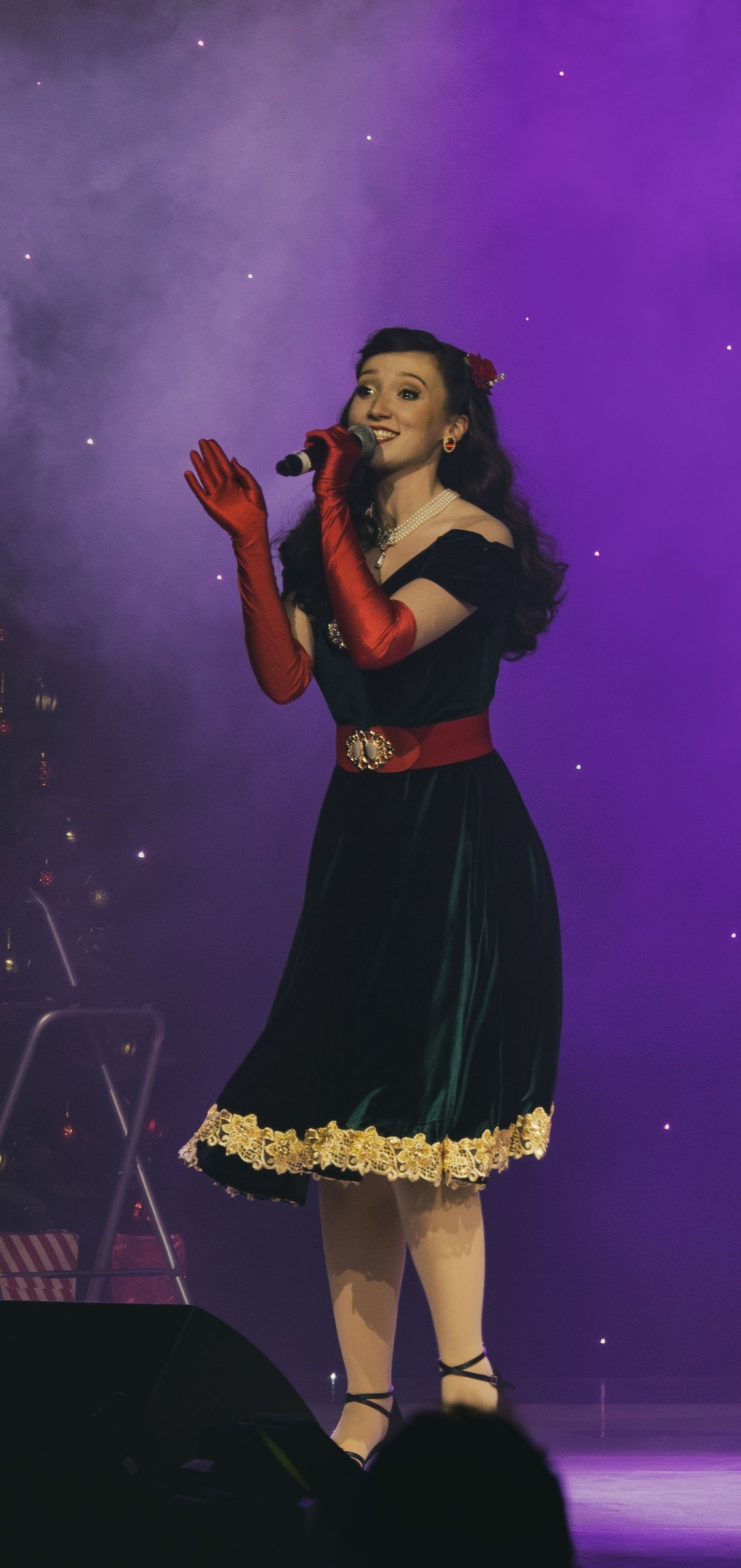 A woman singing on stage with purple lighting, wearing a vintage style black dress with gold lace trim, red gloves, and a red belt with a jewel, holding a microphone.