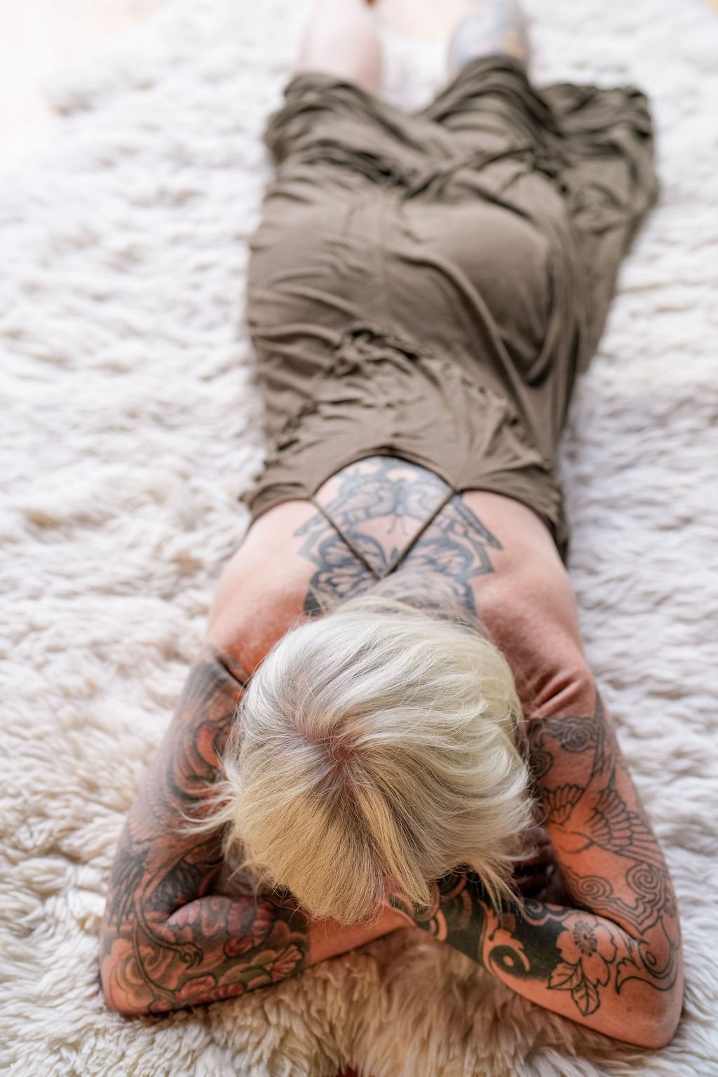A woman with tattoos and platinum blonde hair lying on her stomach on a white fur rug.