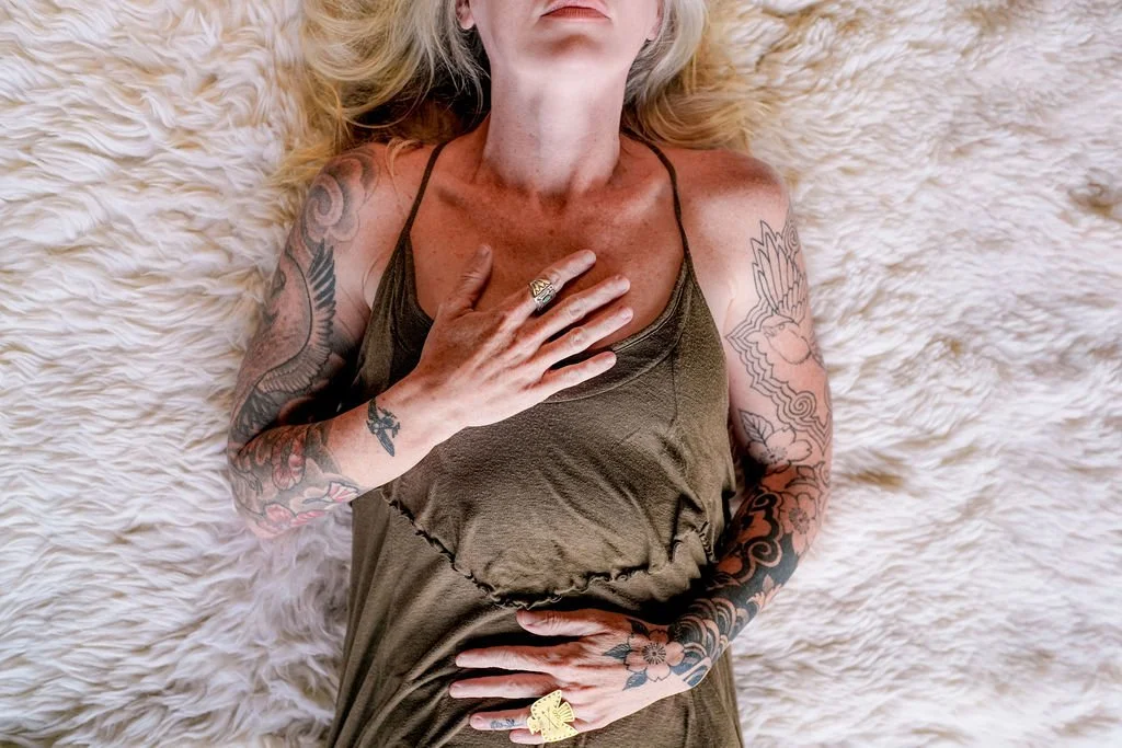 A woman with tattoos lying on a fluffy surface, wearing a brown dress, with her hand on her chest and stomach.