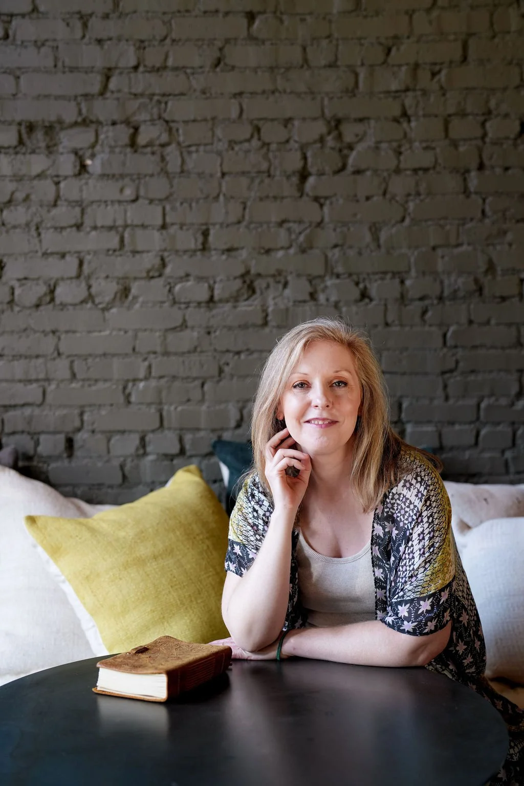 A woman with blonde hair sitting at a round table, resting her chin on her hand, smiling, with a hardcover book in front of her, against a background of a dark brick wall.