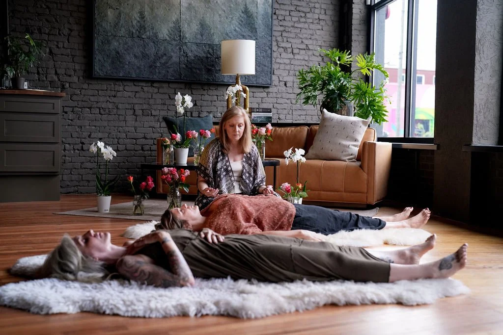 Three women lying on a fluffy white rug on a wooden floor, with the woman in the middle holding a card, in a cozy, decorated room with flowers, a leather couch, and a large window.