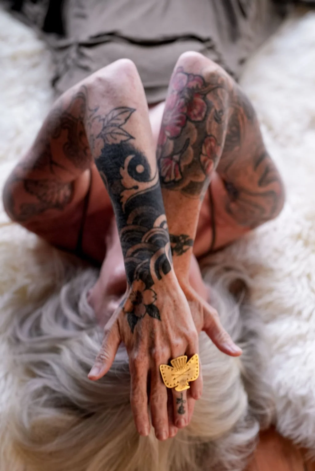 Close-up of tattooed hands with a large gold ring, resting on a fluffy white surface. The person has tattoos on their arms and fingers, including floral and abstract designs.