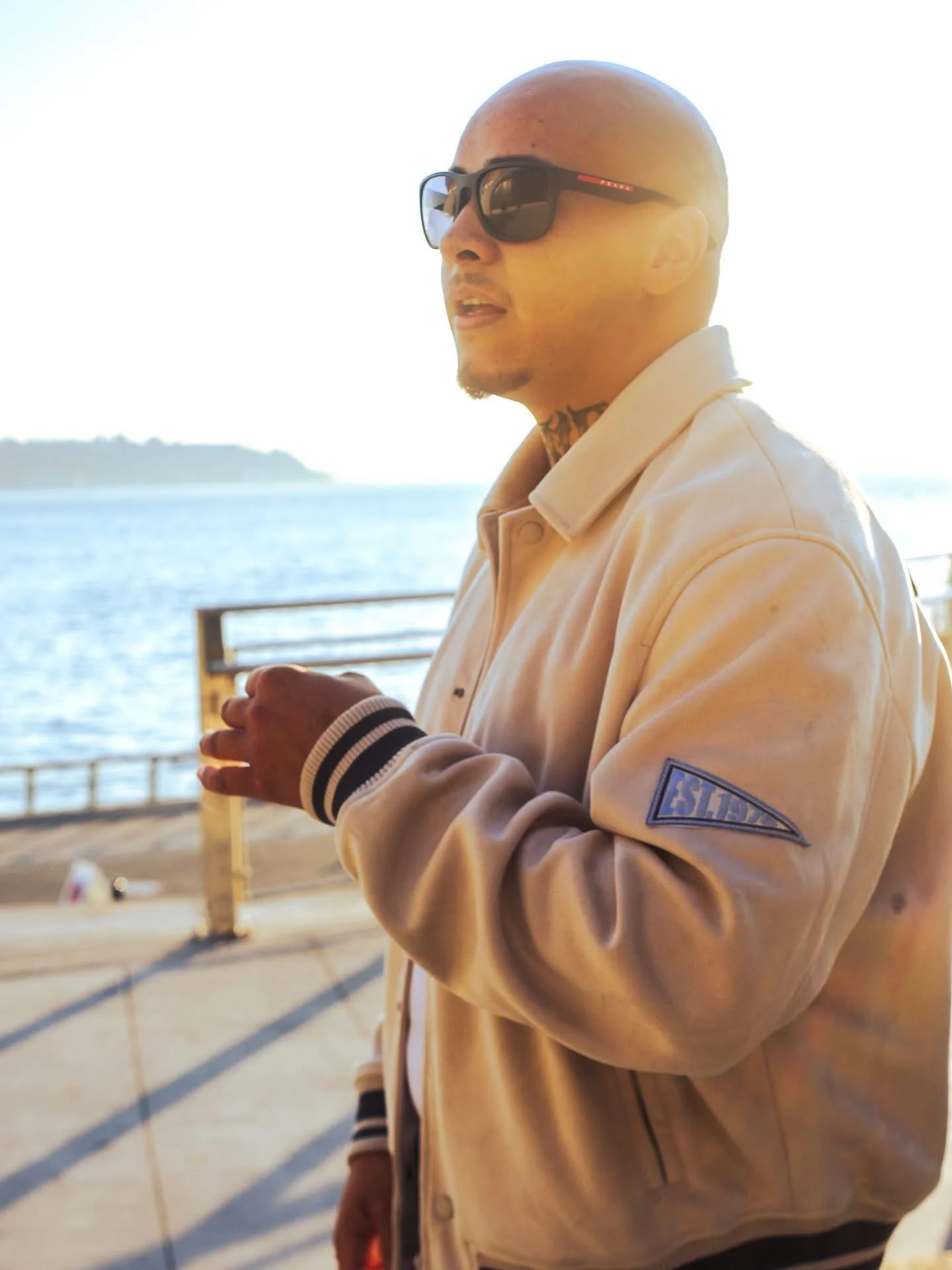 GMDCASH wearing sunglasses and a beige jacket standing near water with a bridge in the background.
