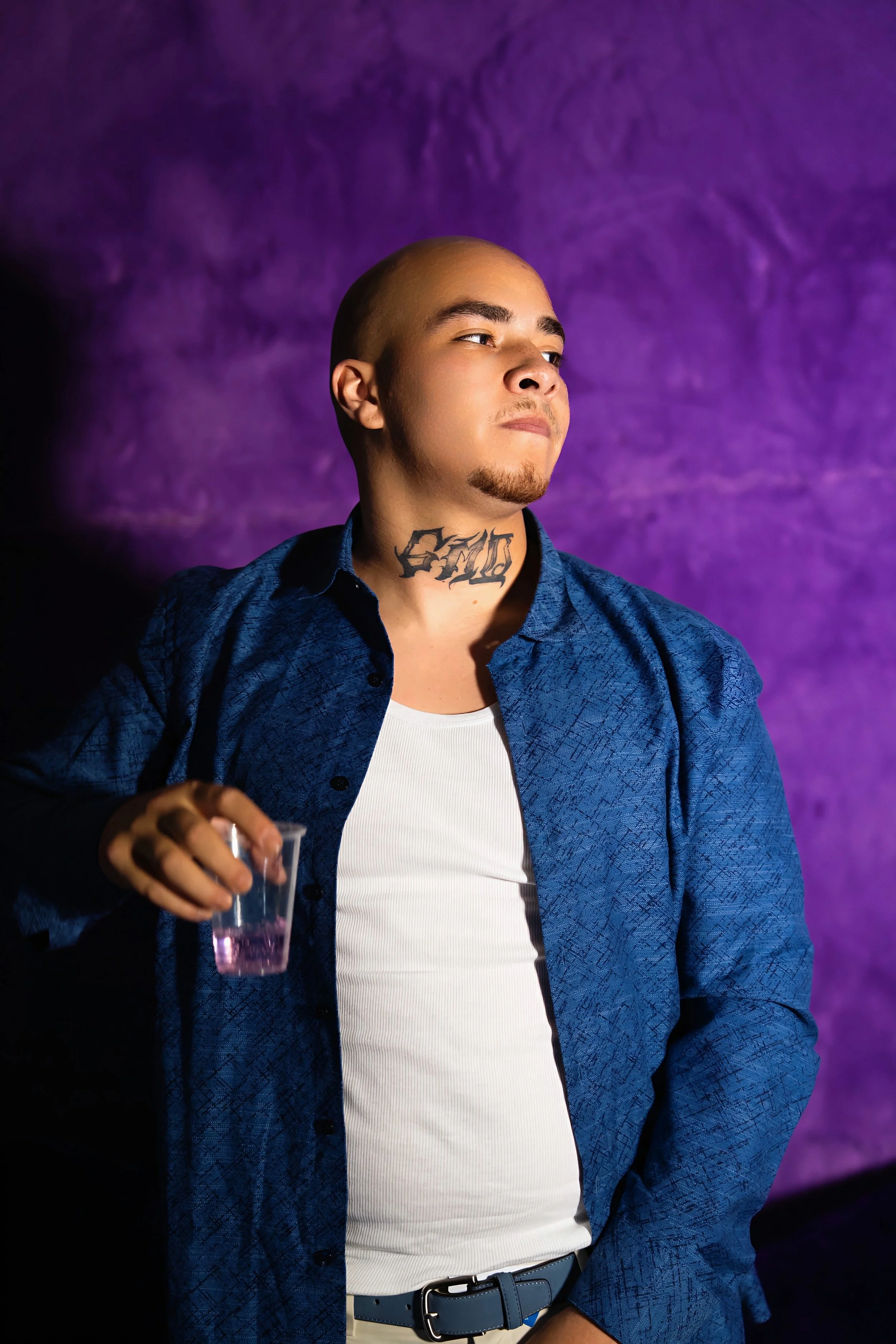 GMDCASH with a tattoo on his neck, wearing a white shirt and blue jacket, holding a clear cup with purple liquid, standing in front of a purple background.