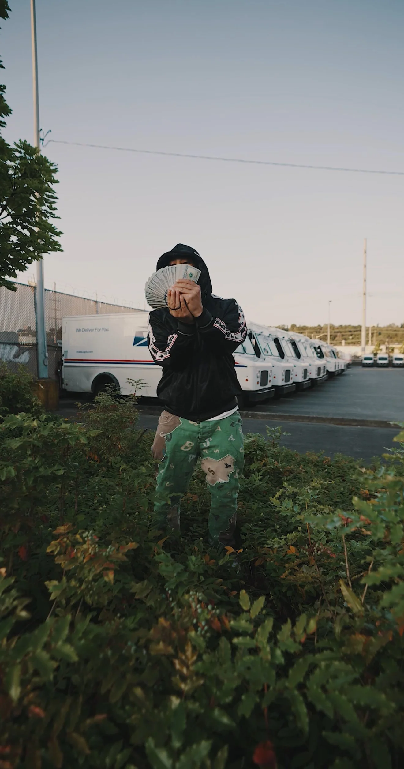 GMDCASH standing outdoors behind bushes, holding a fan of cash in front of their face, with postal trucks and utility poles in the background.