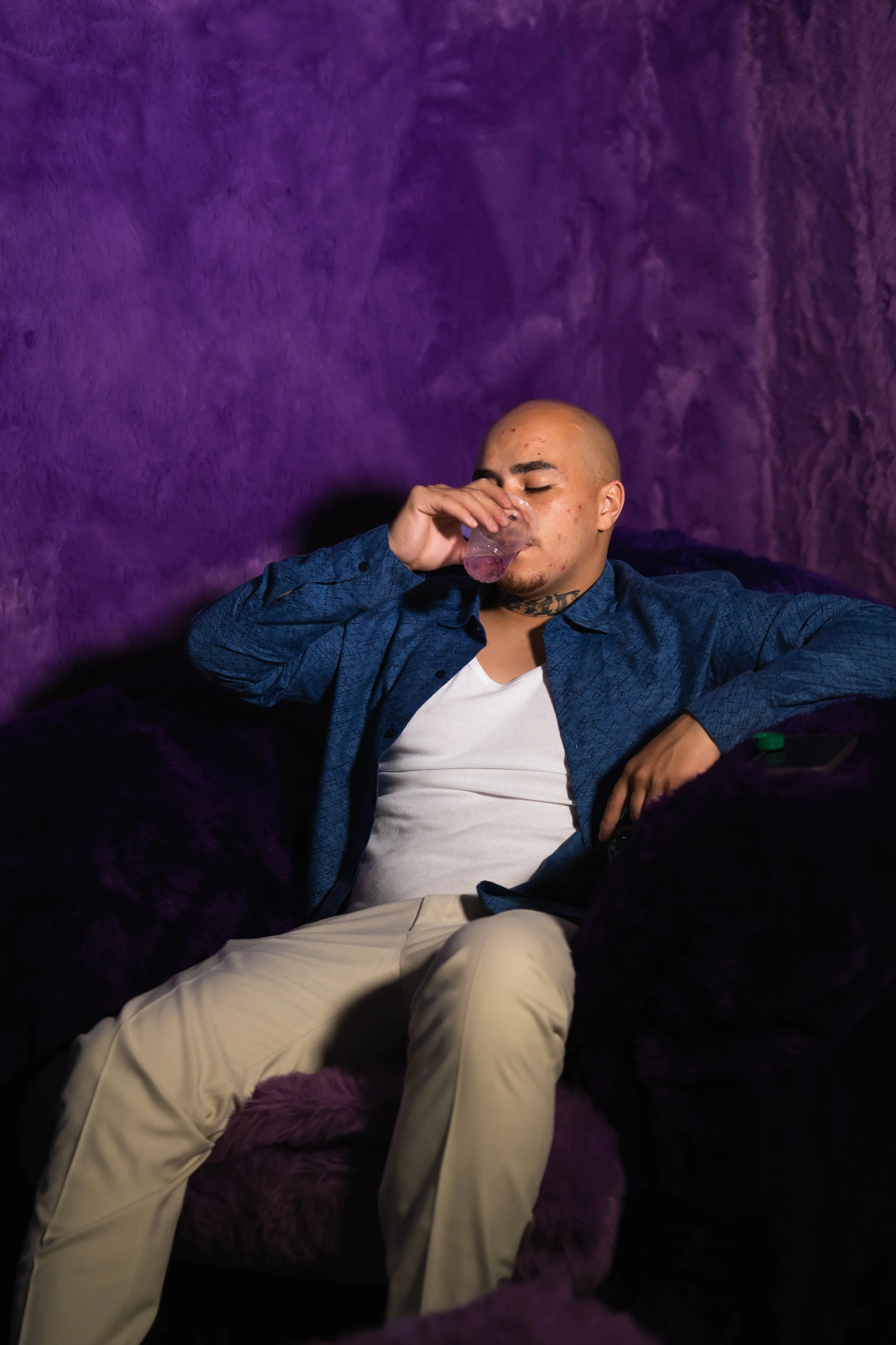 A bald man with tattoos on his neck is sitting on a purple cushioned chair or sofa, wearing a white t-shirt and a blue patterned shirt. He is drinking from a glass with a purple-colored beverage, with a calm or relaxed expression. The background is p