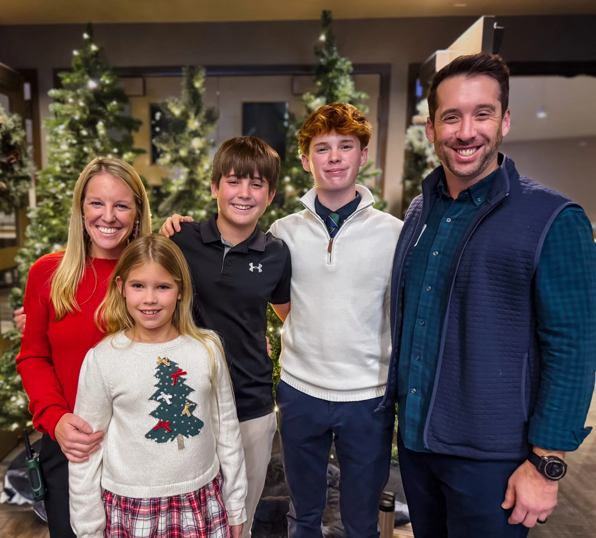 A family of six people, including two women and four men, smiling and standing in front of decorated Christmas trees indoors during the holiday season.