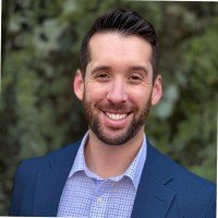 Portrait of a smiling man outdoors, wearing a blue blazer and dress shirt, with a blurred green background.