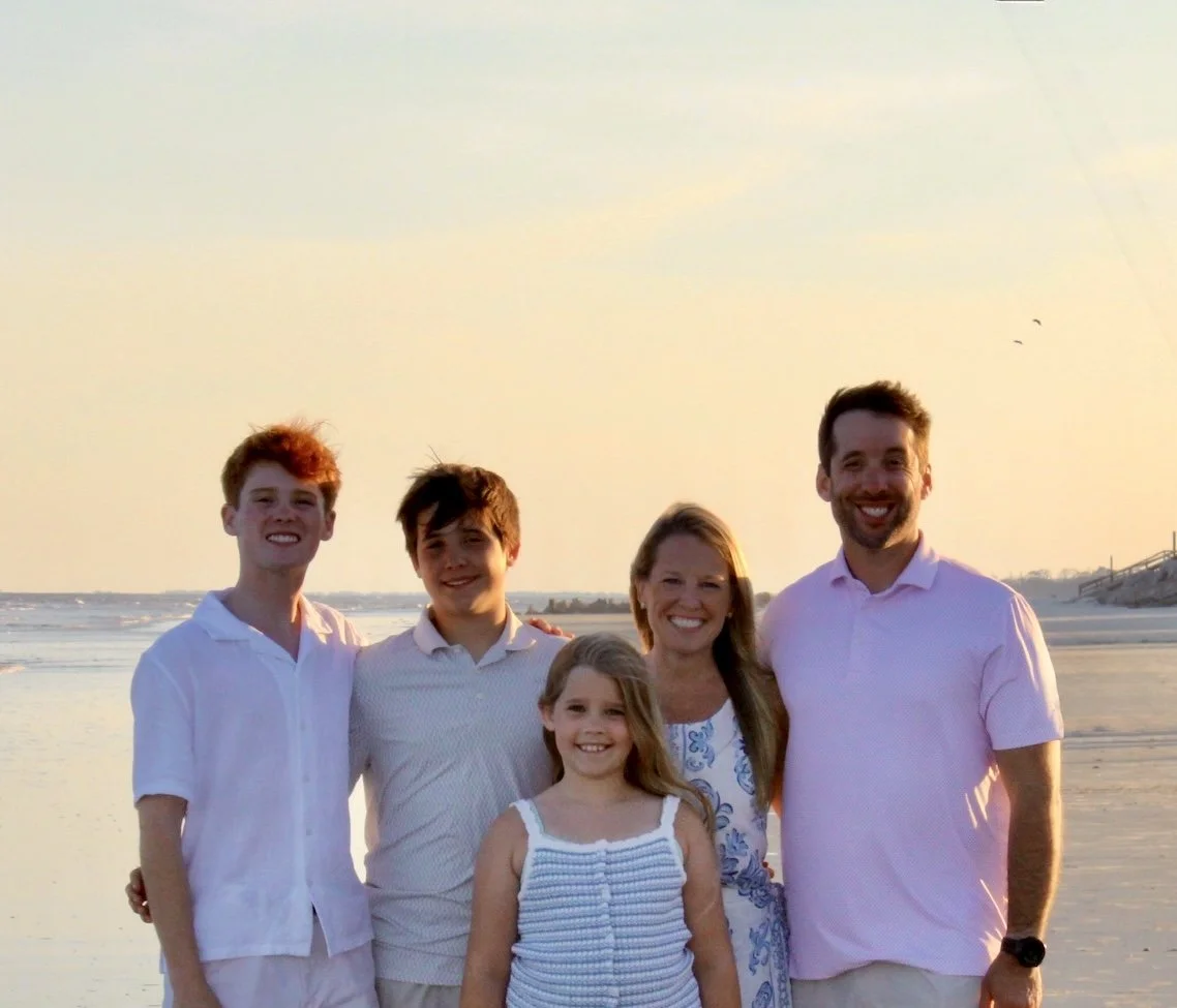 Family of five smiling on the beach during sunset, with water, sand, and a few birds in the background.