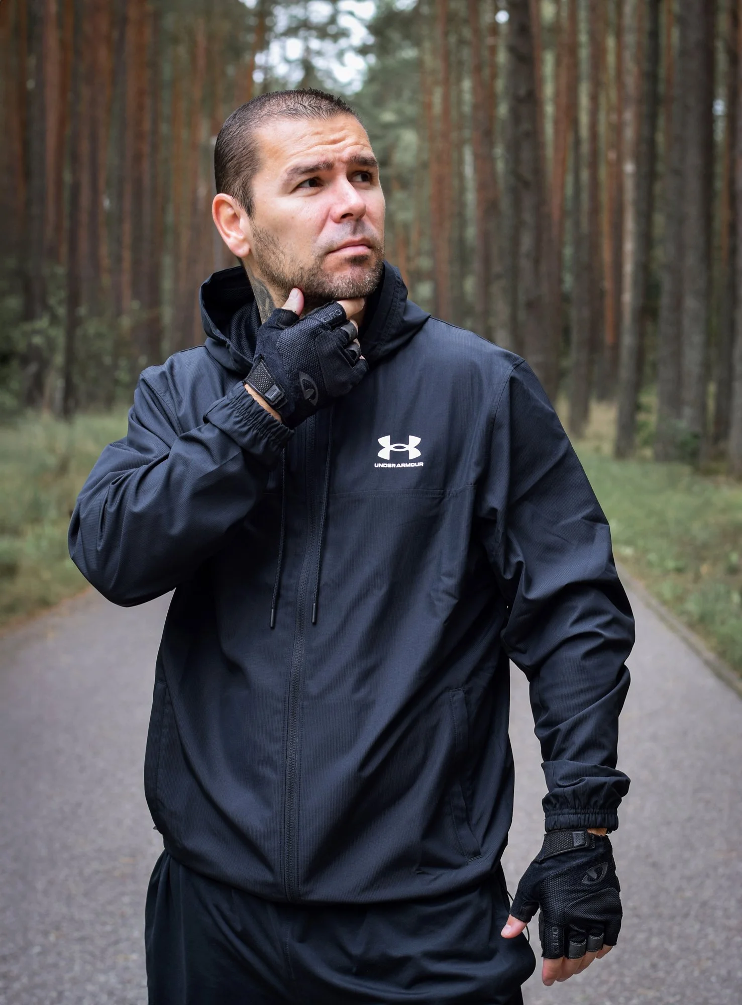 Man in black Under Armour jacket and gloves standing outdoors in a forest with trees in the background, holding his chin with his right hand in a thoughtful pose.