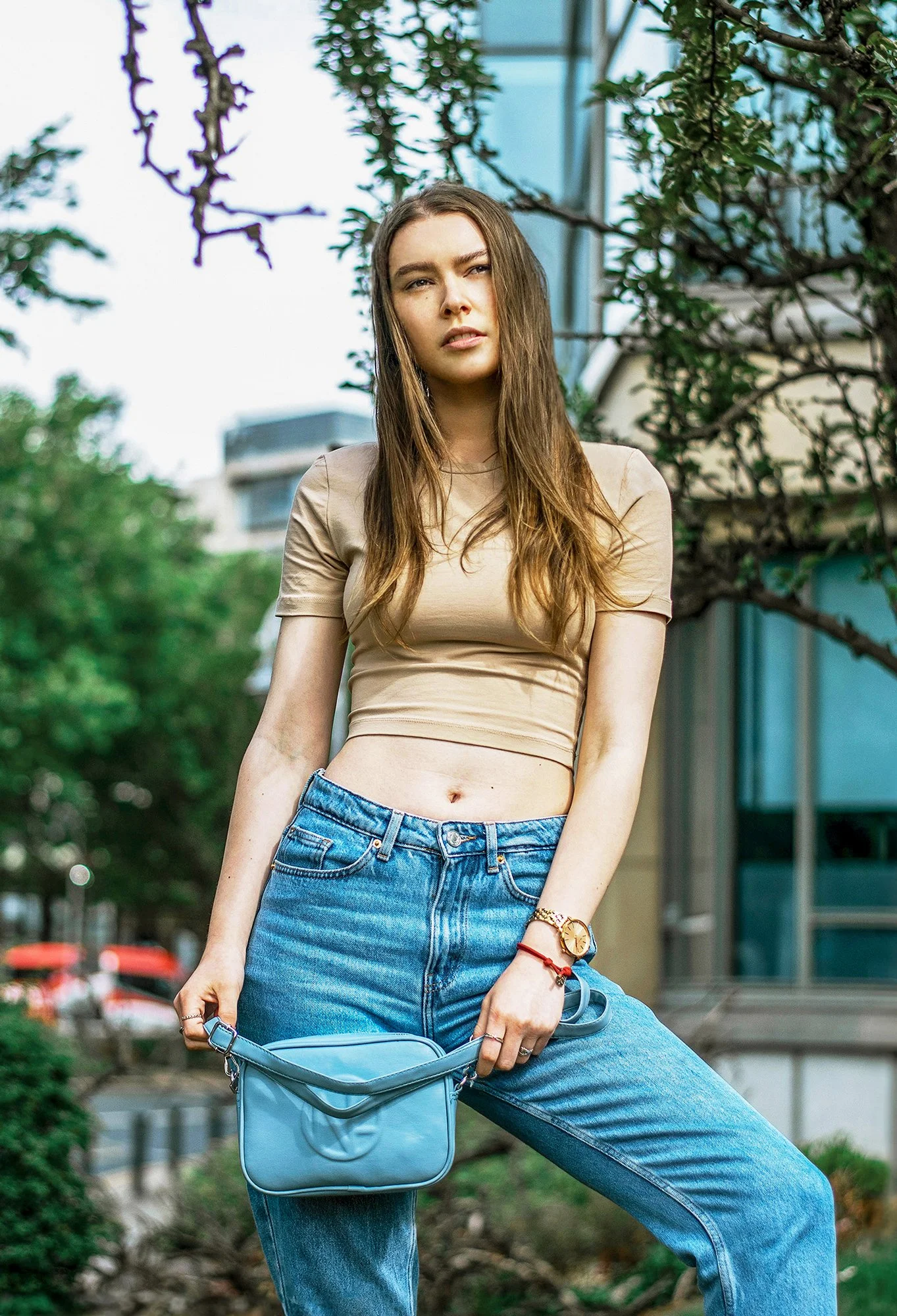 A woman wearing a beige crop top and blue jeans standing outdoors on a city street with trees and modern buildings in the background. She is holding a light blue purse. Lifestyle product photography image showcasing a purse.