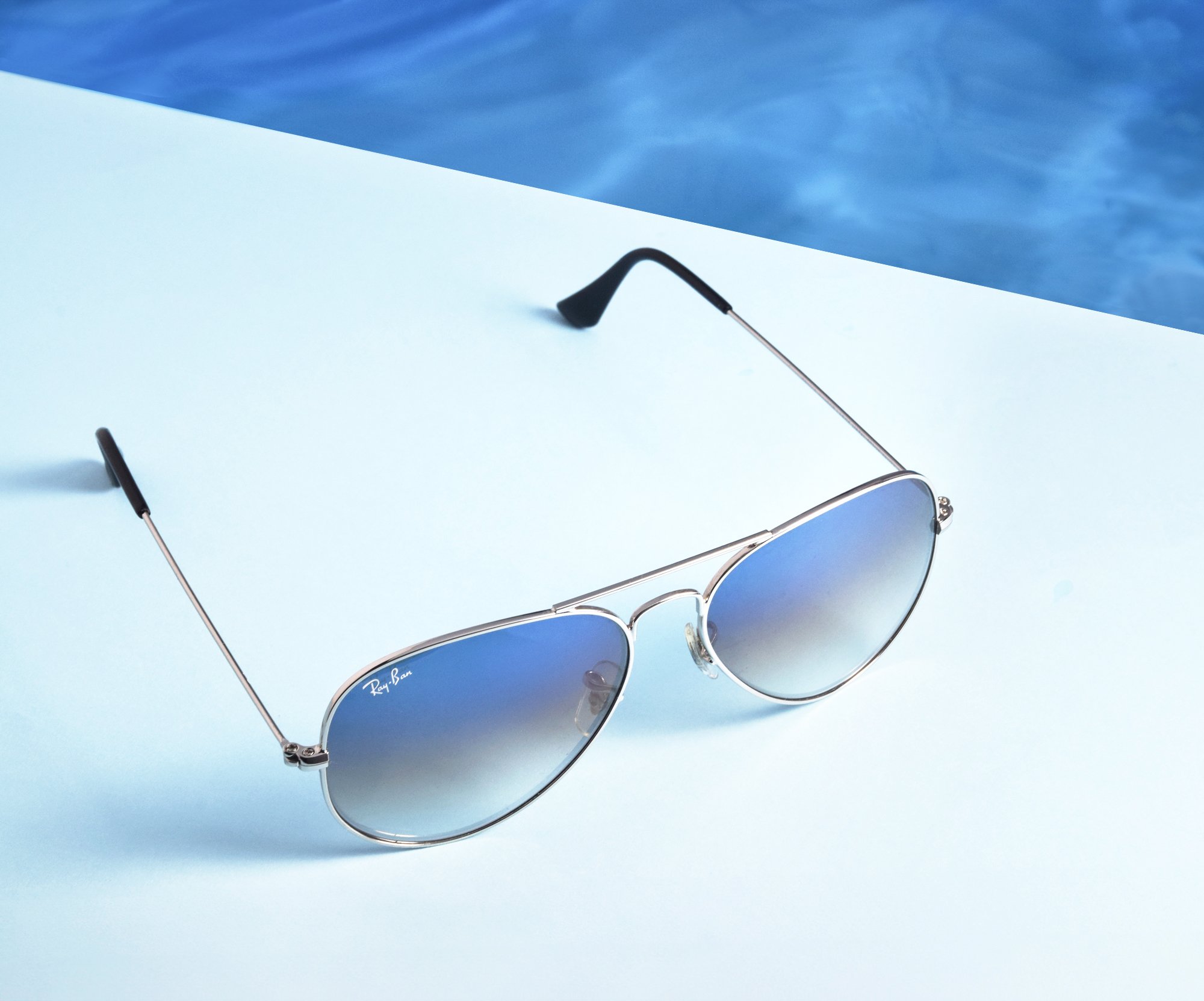 A creative still-life product photo showcasing a pair of sunglasses  with blue tinted lenses resting on a light blue surface, with a portion of a blue swimming pool visible in the background.