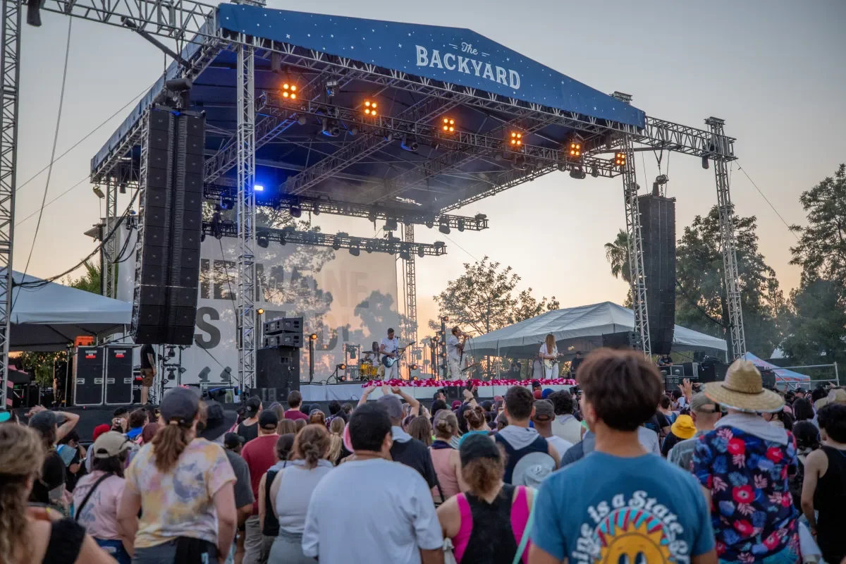 An outdoor concert with a band performing on a large stage at dusk, crowd of people watching, some wearing hats and colorful clothing.