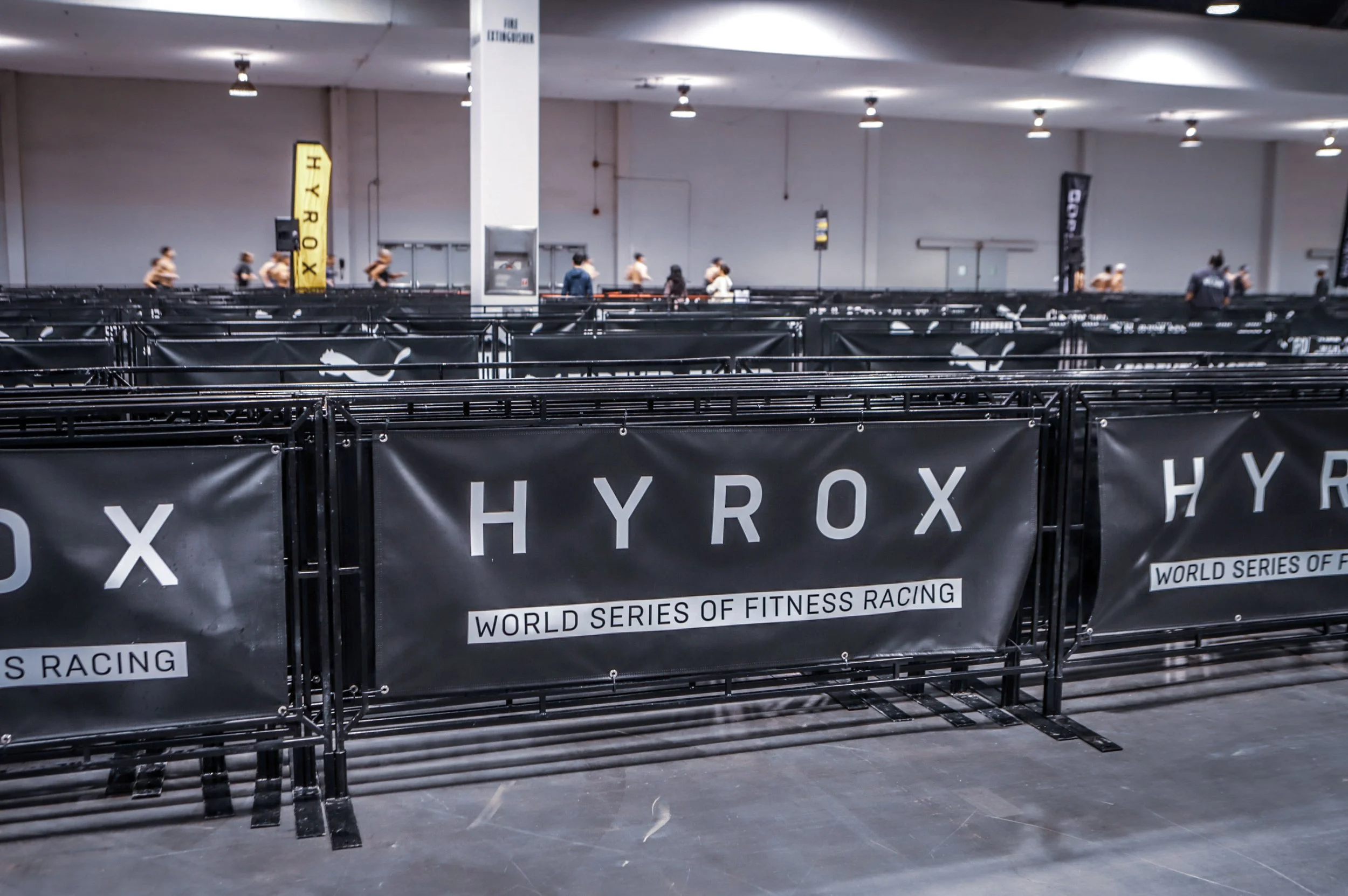 Indoor fitness racing venue with black barriers displaying 'HYROX' and 'World Series of Fitness Racing' banners, and participants in athletic gear training in the background.