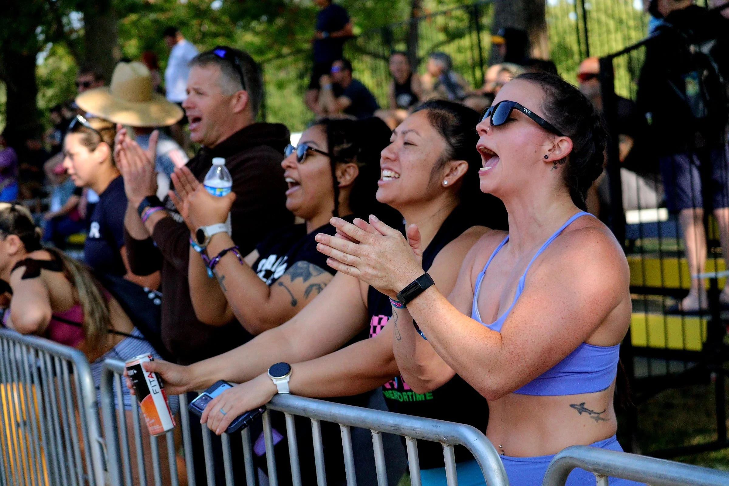 Group of people at a concert or outdoor event, cheering and singing along, standing behind a metal barricade, with trees in the background.