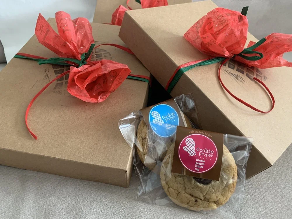Two gift boxes wrapped in kraft paper with red tissue paper bows and green and red ribbon. Two cookies in clear plastic packaging with stickers that say 'Cookie Proper' and describe the flavors.