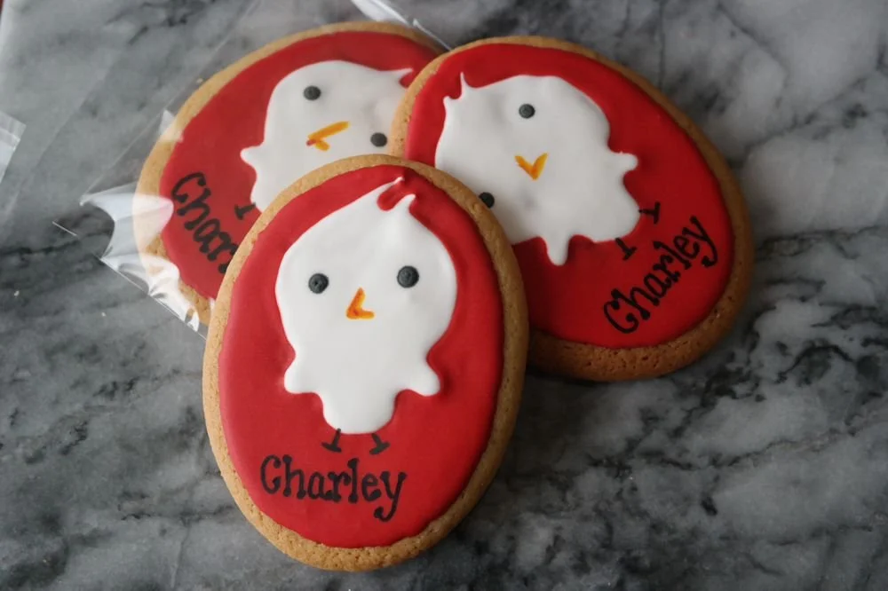 Oval sugar cookies decorated with red icing and a white ghost face with black eyes and an orange beak, resembling a chick. The cookies have the name "Charley" written in black icing.