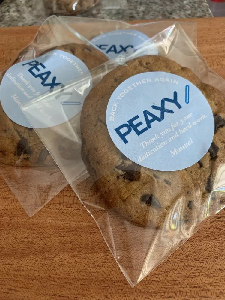 Three chocolate chip cookies in clear plastic bags with blue personalized labels that say "BACK TOGETHER AGAIN PEAX! Thank you for your dedication and hard work. Manuel." on a wooden surface.