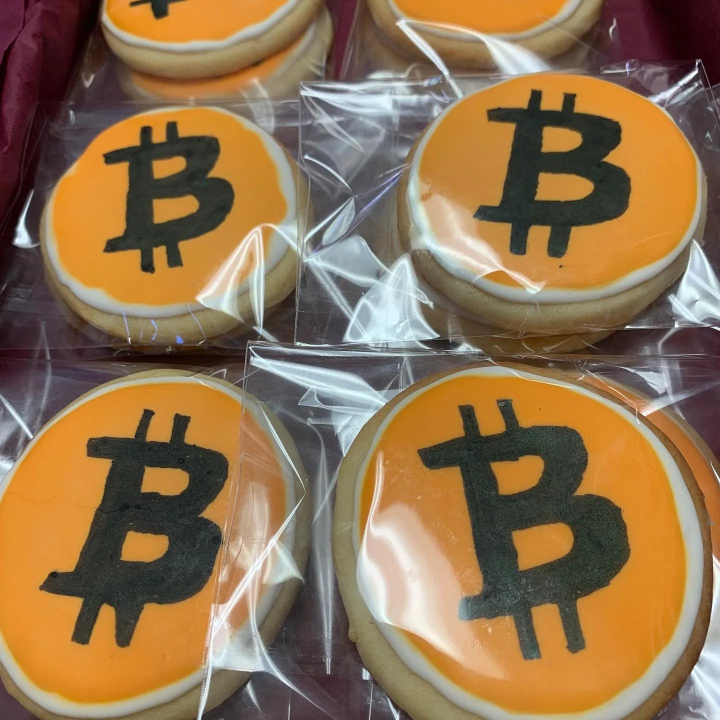 Cookies decorated with the Bitcoin symbol in black on an orange background, individually wrapped in clear plastic.