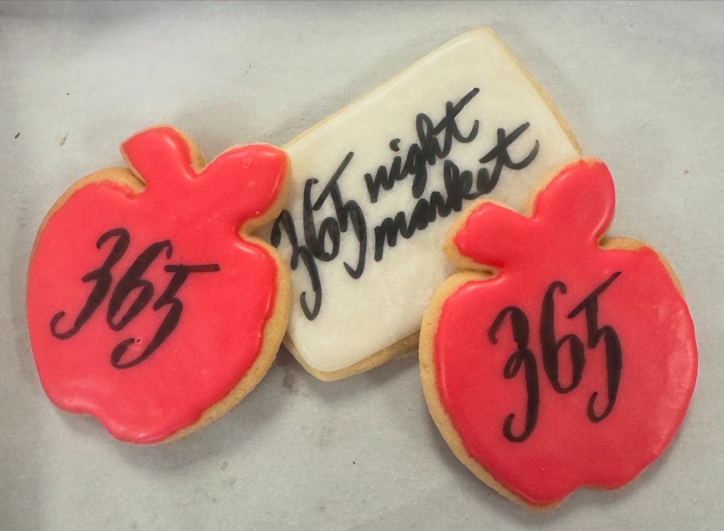 Decorated cookies with the number 365 written on peach-colored apple shapes and a rectangular white cookie with the handwritten message '365 nights of heartbreak' in black icing.
