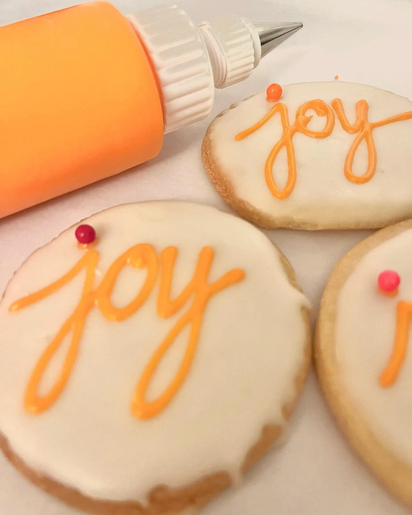 Decorated sugar cookies with the word "joy" written in orange icing on top, and colorful small round candies on each cookie. A large orange and white icing piping bag is in the background.