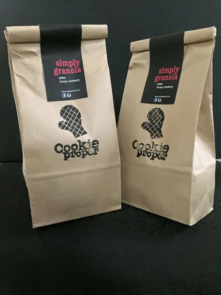 Two brown paper bags with black labels and logo, labeled 'Simply Granola' with flavor 'paleo honey cranberry' from Cookie Prop.