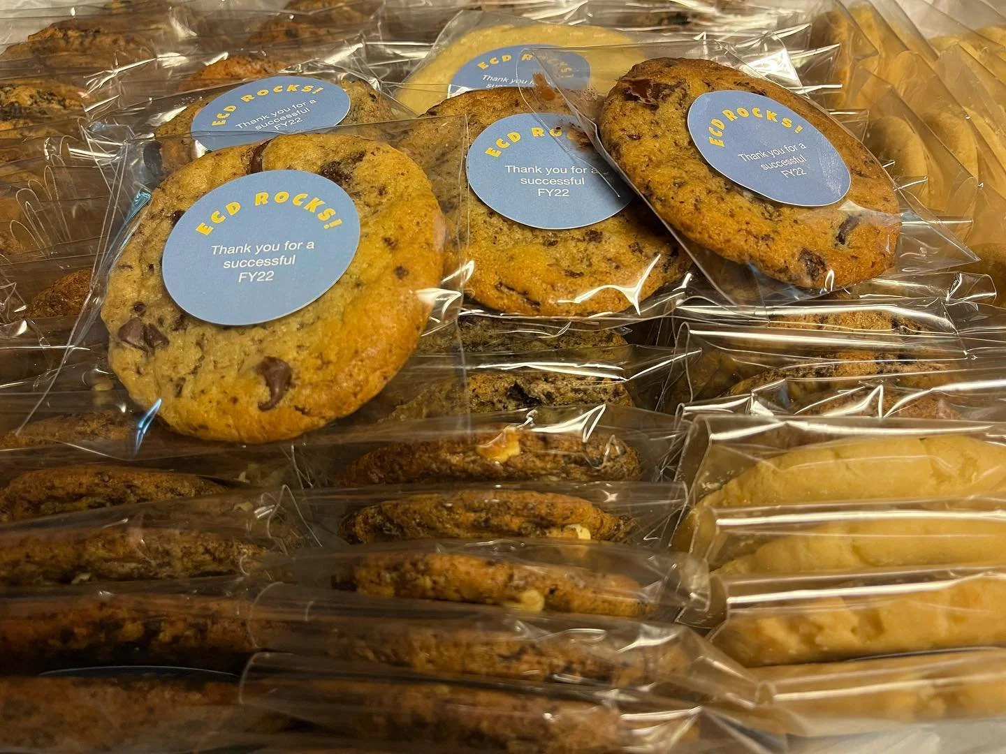 A tray of individually wrapped cookies, including chocolate chip and possibly peanut butter cookies, with some labeled with a 'Thank you for a successful FY22' sticker.