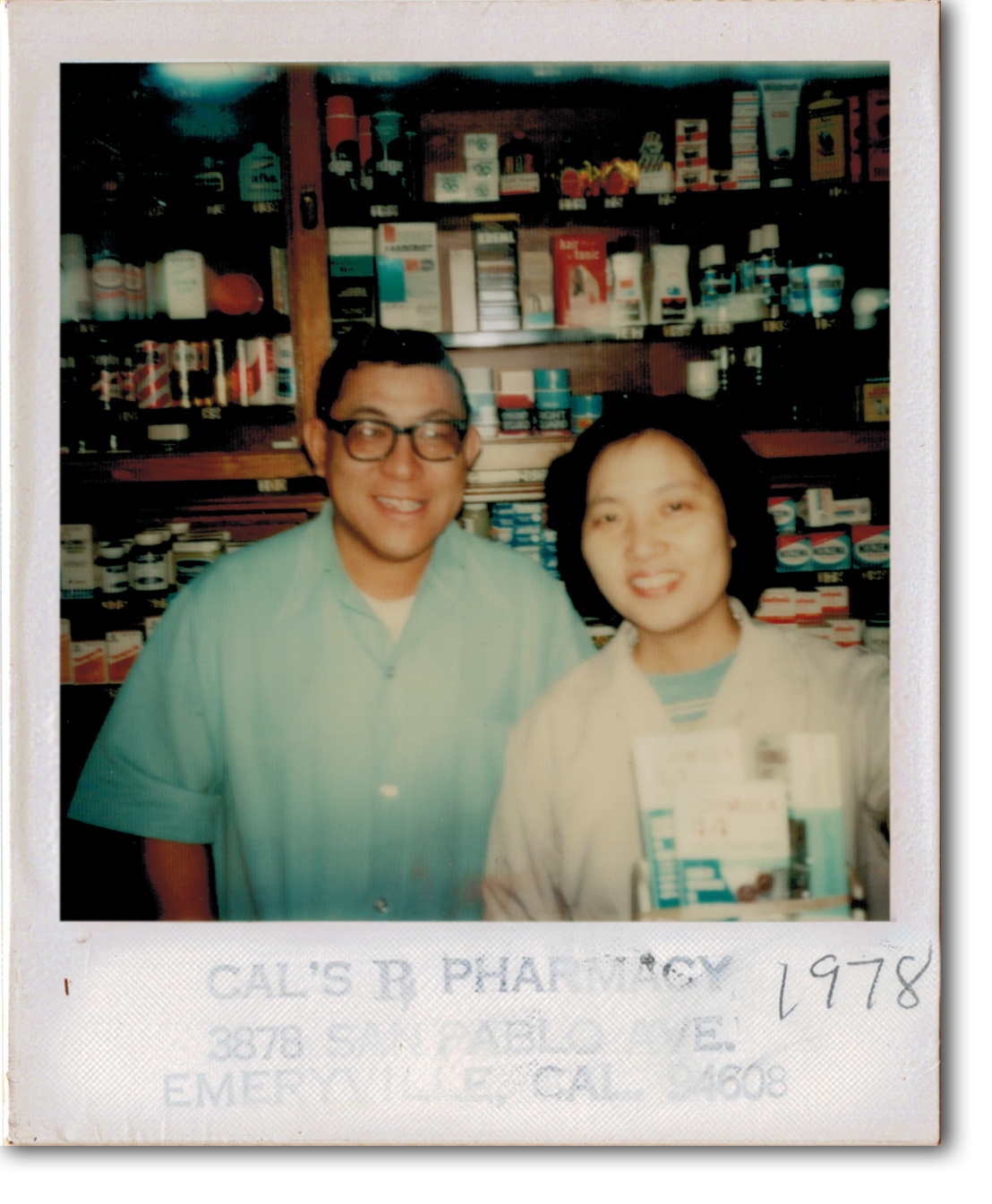 A man and a woman smiling for a selfie in a pharmacy, with shelves of health products and medicines behind them. The photo appears to be from 1978 and was taken at Cal's Pharmacy, located at 2878 S. Tranel, E. Emeryville, California.