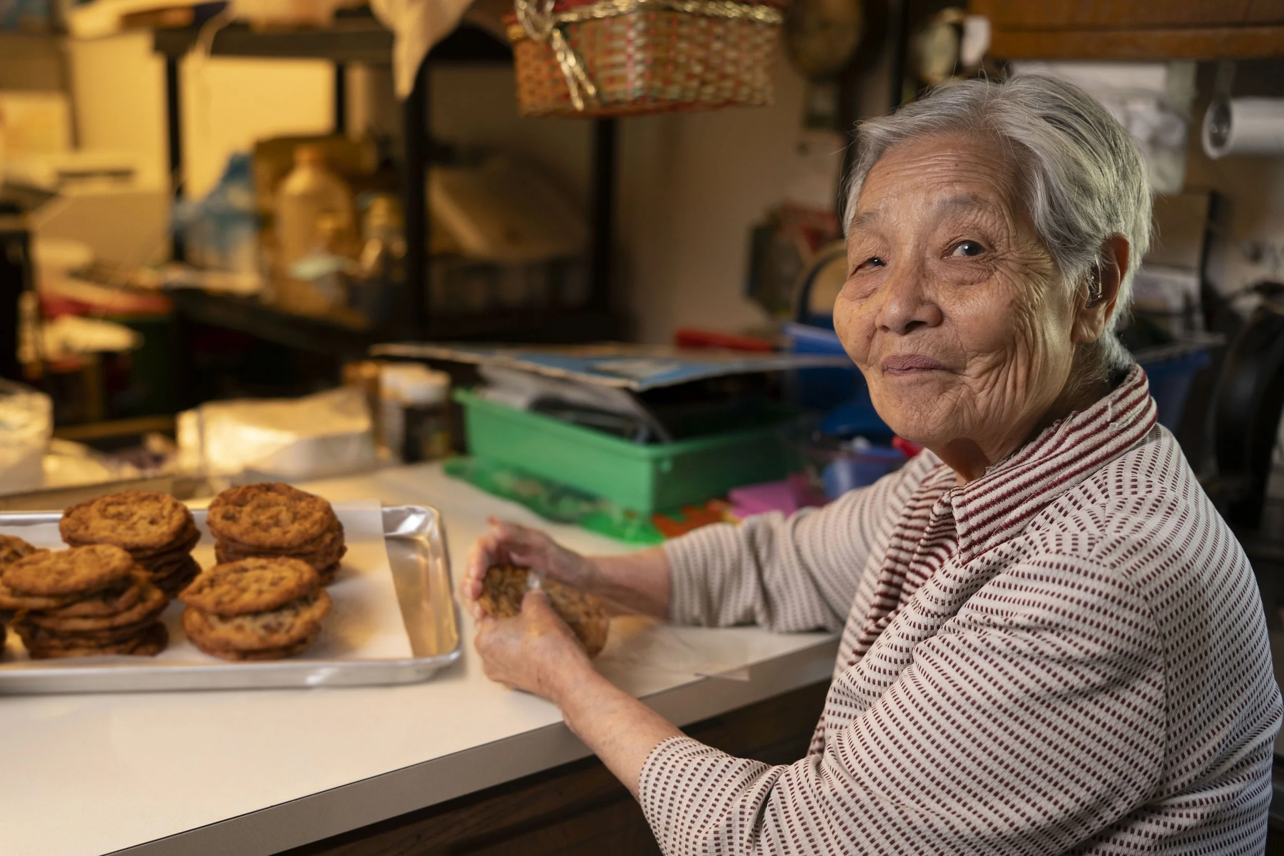 An elderly woman with gray hair smiling while sitting at a counter with a tray of cookies nearby, in a cozy kitchen or bakery setting.