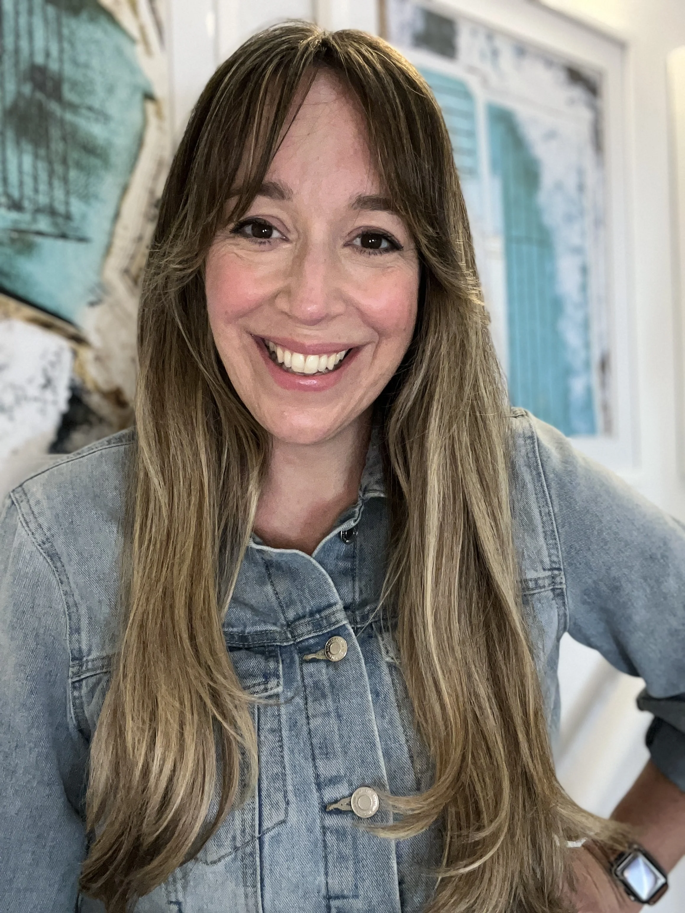A woman with long hair smiling at the camera, wearing a denim jacket, standing in front of ocean background art
