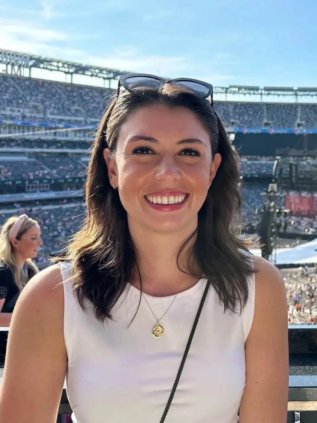 A woman wearing a white sleeveless top and sunglasses on her head, smiling at the camera at a sports stadium during the day with a large crowd in the background.