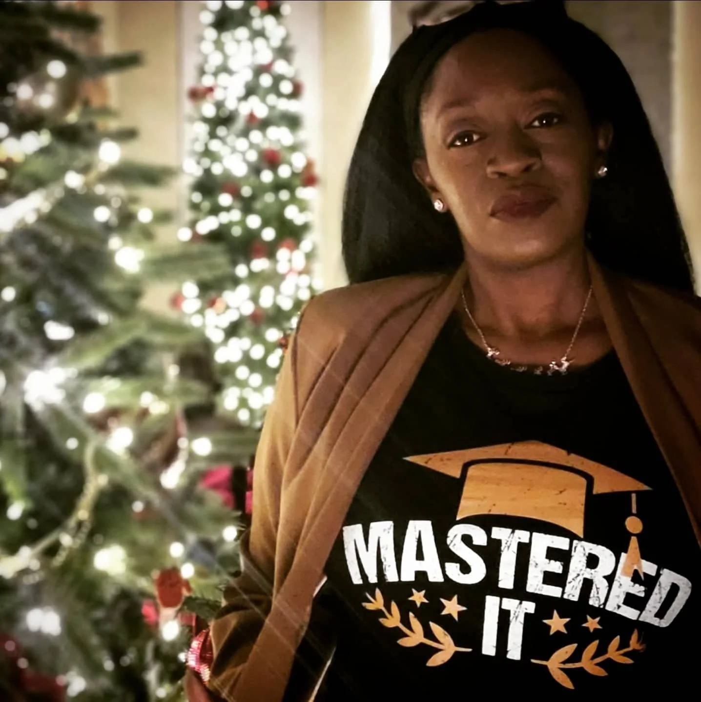 Portrait of a professional woman standing indoors near a decorated Christmas tree, wearing a blazer and a “Mastered It” shirt.