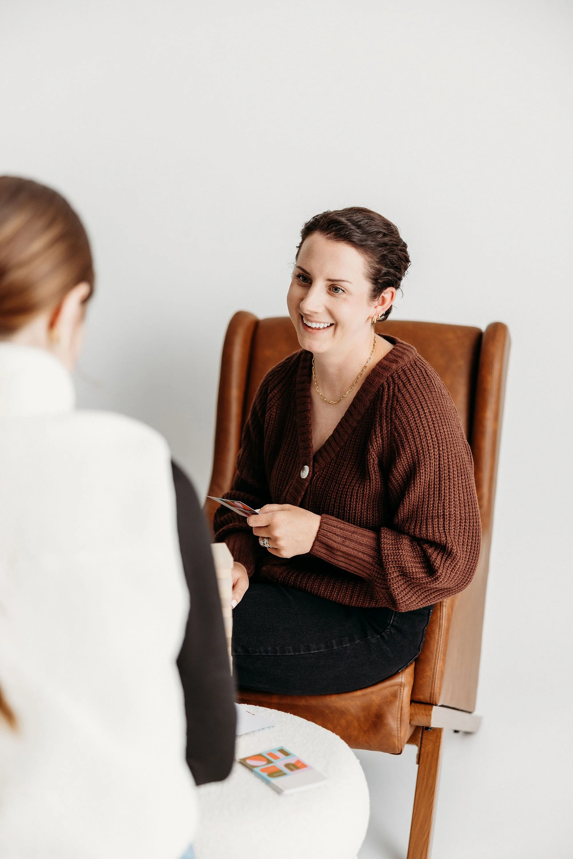 A woman with short dark hair and hoop earrings is smiling and talking to another person while sitting on a wooden chair. There is a white table with some colorful cards on it.