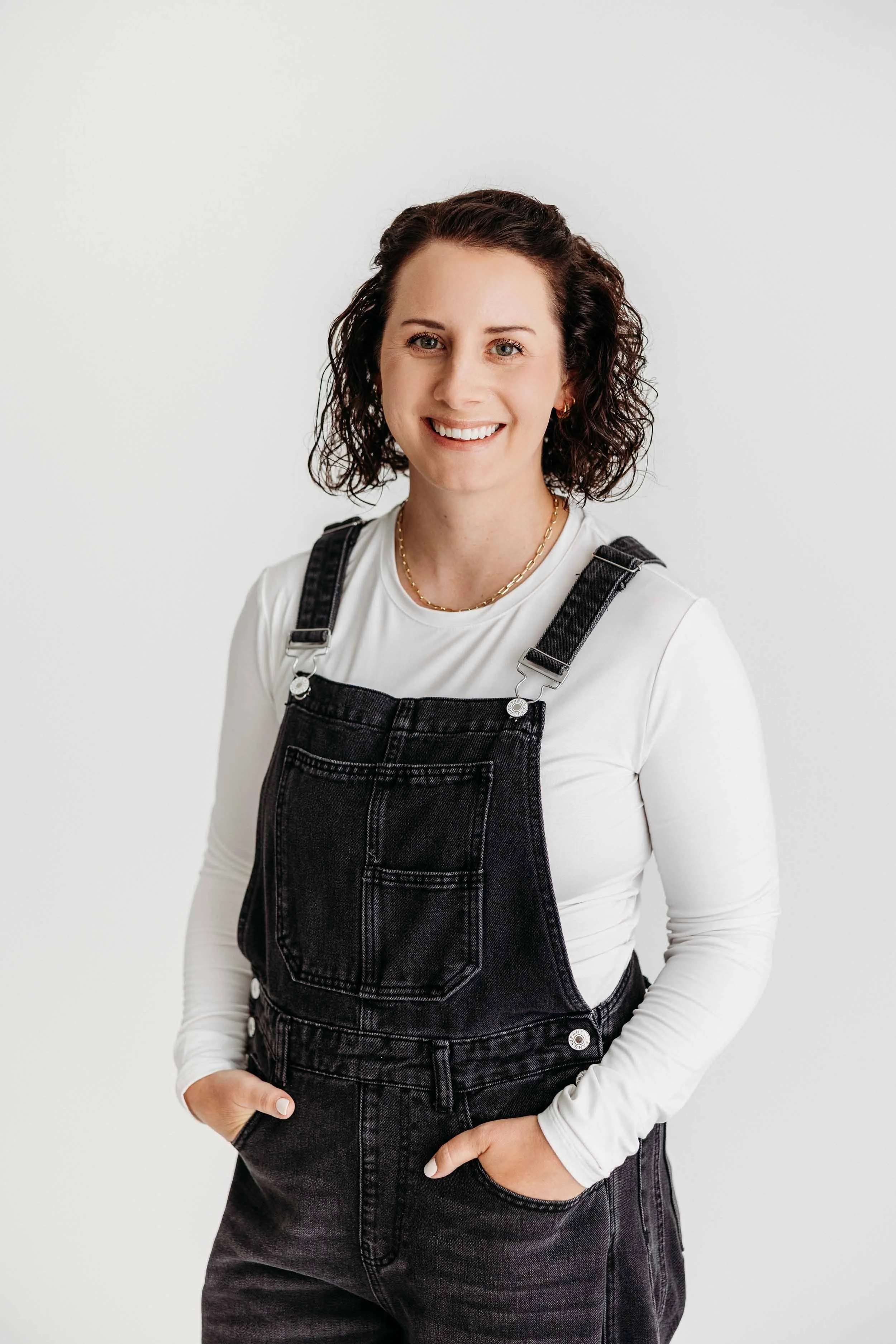 A woman with curly brown hair smiling, wearing a white long sleeve shirt, black denim overalls, and a gold chain necklace, standing against a plain white background.