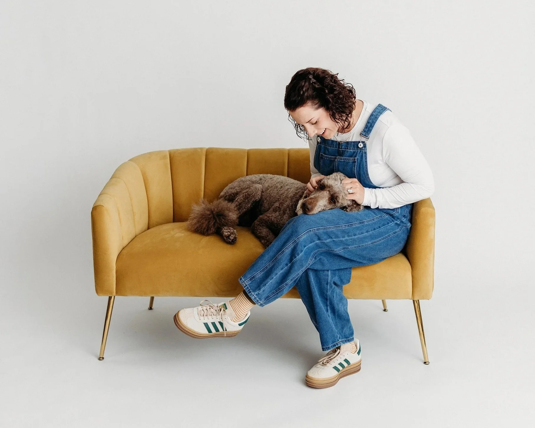 A woman sitting on a yellow mid-century modern sofa with metal legs, leaning forward with a smile while petting a brown poodle laying on the sofa. She is wearing a white long-sleeve shirt, denim overalls, and white sneakers with green stripes, with her feet crossed at the ankles. The background is plain white.
