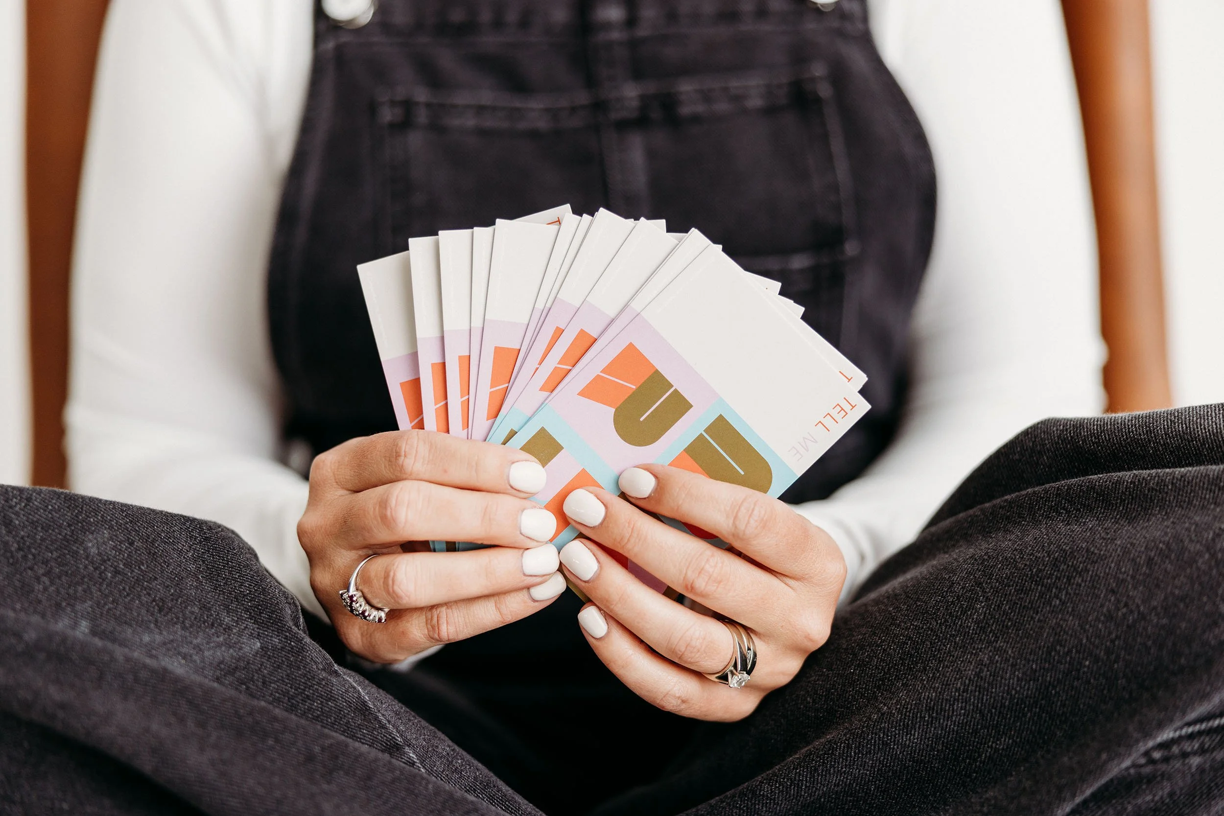Person holding a fan of colorful cards or tickets, wearing dark jeans and a white long-sleeve shirt.