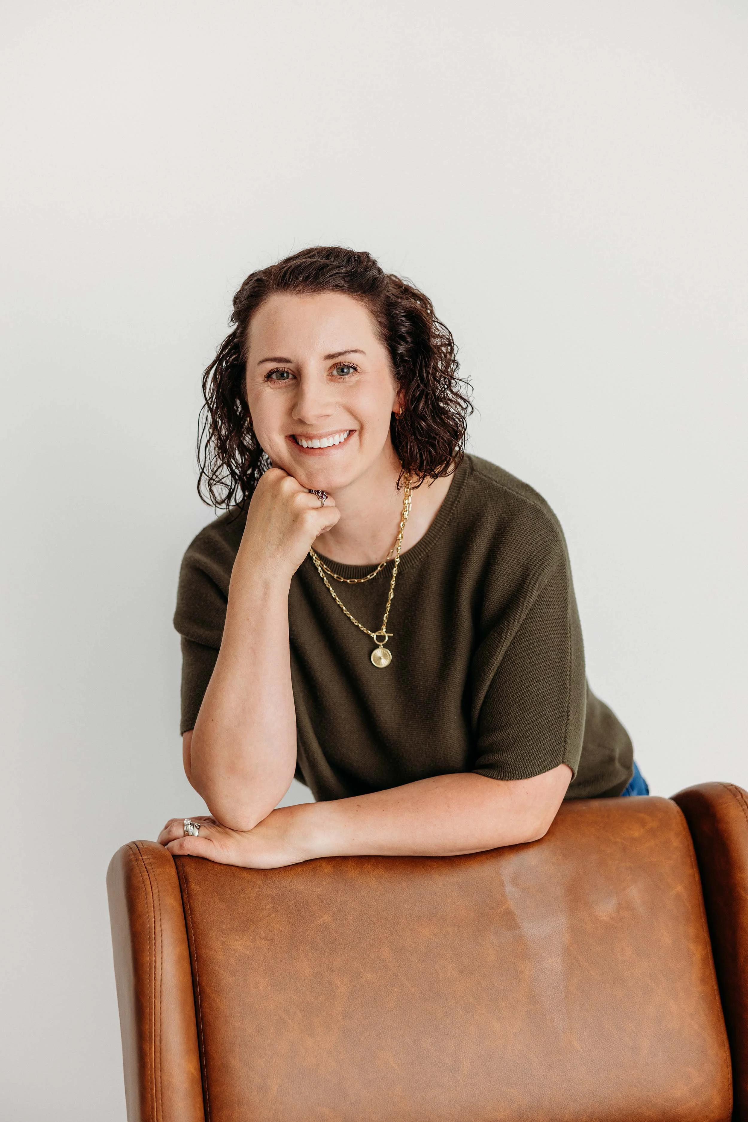A woman with curly brown hair smiling, resting her chin on her hand, wearing a green sweater and gold jewelry, sitting in front of a plain white wall.