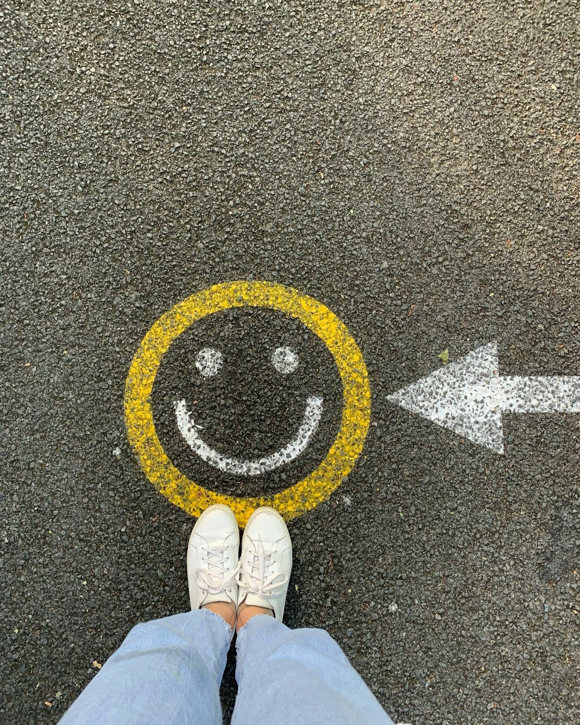 A person's feet wearing white sneakers and light blue pants standing on asphalt with a yellow circle and a white arrow painted on the ground.