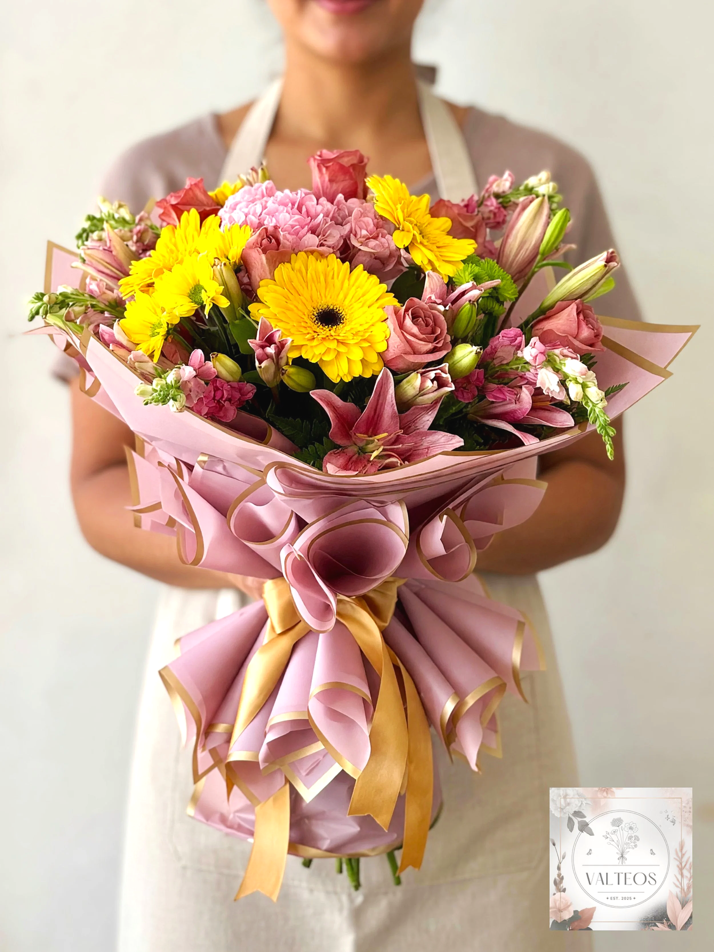 Valteos Flowers and Gifts. A woman holding a bouquet of pink, yellow, and green flowers wrapped in pink paper with gold accents.