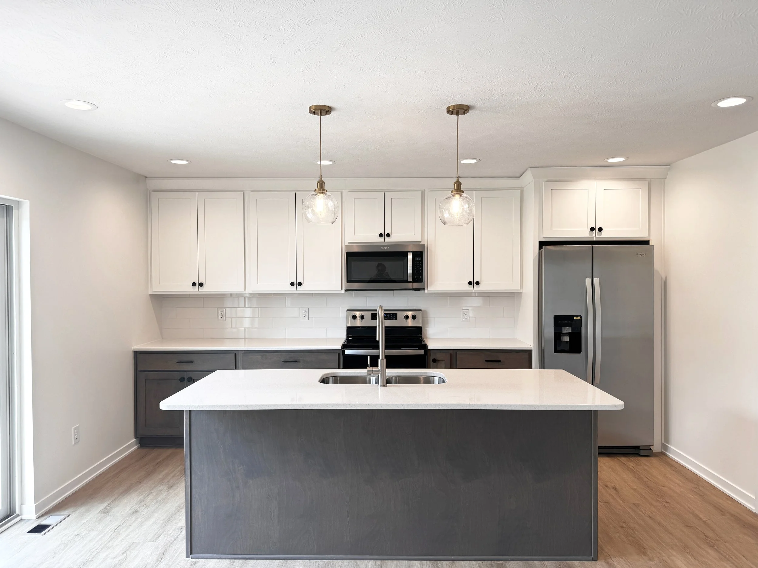 Modern kitchen with white upper cabinets, gray lower cabinets, a kitchen island with a white countertop, stainless steel refrigerator, microwave, and stove, pendant lights, and hardwood flooring.